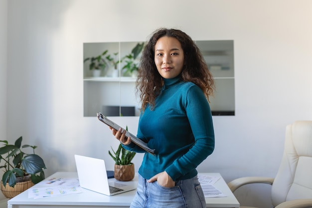 Lächelnde junge asiatische Mitarbeiterin steht am Schreibtisch im Büro und blickt in die Ferne, denkt oder visualisiert den beruflichen Erfolg Glückliche Geschäftsfrau plant oder träumt am Arbeitsplatz Business-Vision-Konzept