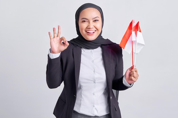 Lächelnde junge asiatische muslimische Frau, die eine indonesische Flagge hält und den indonesischen Unabhängigkeitstag feiert, zeigt eine okay-Geste isoliert auf weißem Hintergrund