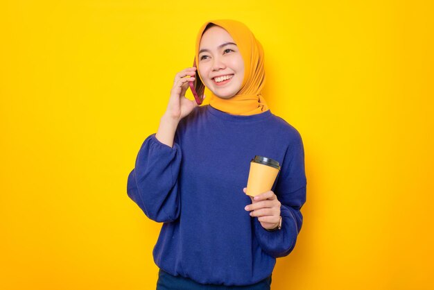 Lächelnde junge asiatische muslimische Frau in einem lässigen Pullover, die auf dem Handy spricht und eine Kaffeetasse aus Papier isoliert über gelbem Hintergrund hält