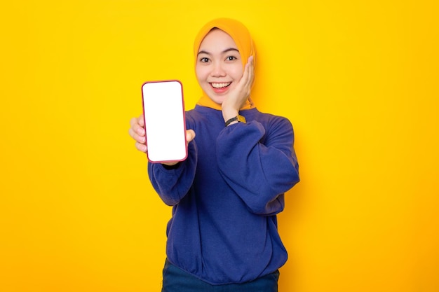 Lächelnde junge asiatische muslimische Frau in einem lässigen Pullover mit einem Handy mit leerem Bildschirm, das eine mobile App isoliert auf gelbem Hintergrund empfiehlt