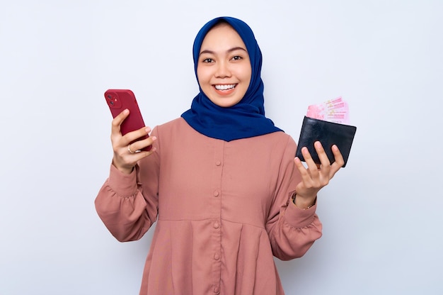 Lächelnde junge asiatische muslimische Frau in rosafarbenem Hemd, die ein Mobiltelefon hält und eine Brieftasche zeigt, die über weißem Hintergrund isoliert ist. Religiöses Lifestyle-Konzept der Menschen