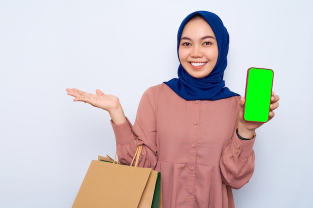 Lächelnde junge asiatische muslimische Frau in rosafarbenem Hemd, die nach dem Einkauf Pakettüten mit Einkäufen hält, die ein Handy mit leerem Bildschirm zeigt und mit den Händen isoliert auf weißem Hintergrund Kopienraum zeigt
