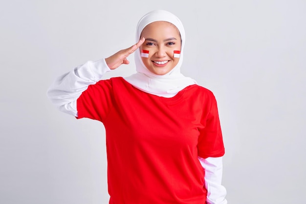 Lächelnde junge asiatische muslimische Frau in rot-weißem T-Shirt, die am 17. August den indonesischen Unabhängigkeitstag feiert und Respektgeste isoliert auf weißem Hintergrund zeigt