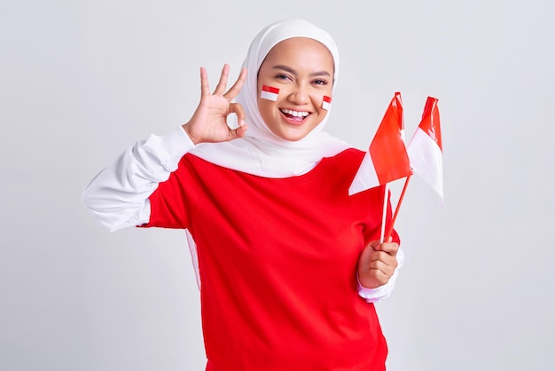 Lächelnde junge asiatische muslimische Frau in rot-weißem T-Shirt, die die indonesische Flagge hält, um den indonesischen Unabhängigkeitstag am 17. August zu feiern und eine okay-Geste isoliert auf weißem Hintergrund zu zeigen