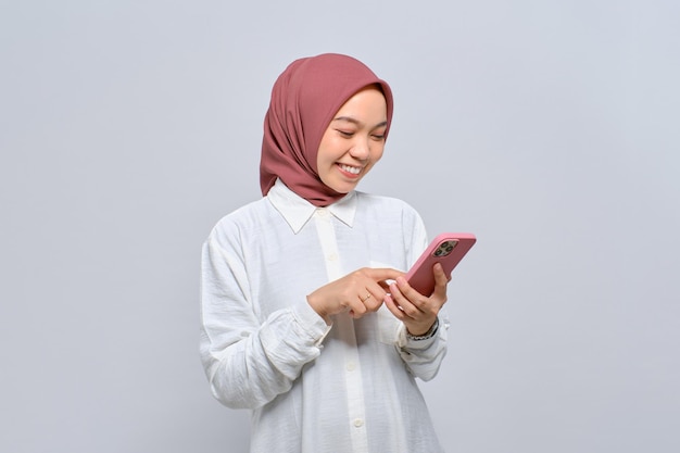 Lächelnde junge asiatische muslimische Frau mit Handy, die gute Nachrichten isoliert auf weißem Hintergrund erhält