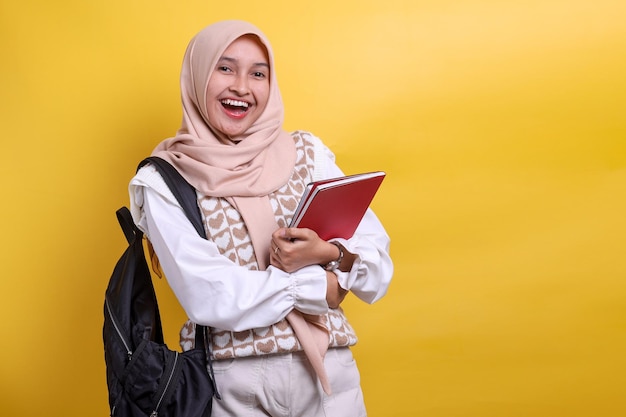 Lächelnde junge asiatische muslimische Studentin im Hijab, die einen Rucksack trägt und Bücher in der Hand hält und in die Kamera blickt