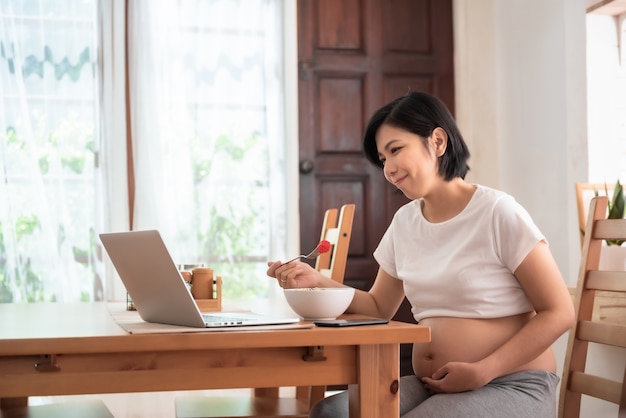 Lächelnde junge asiatische schwangere Frau, die Videoanruf auf Laptop im Esszimmer macht.