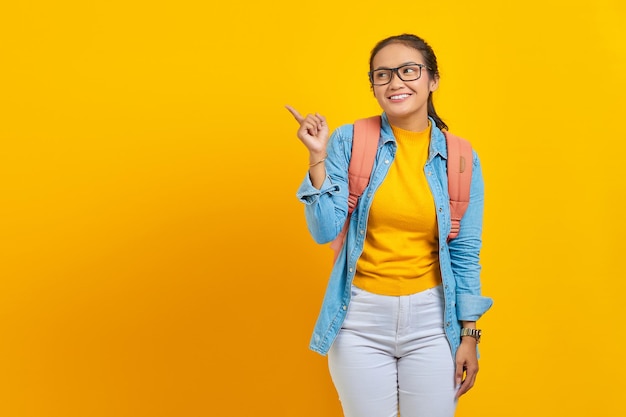 Lächelnde junge asiatische Studentin im Denim-Outfit mit Rucksack, der mit dem Finger in den Kopierraum zeigt und Werbeprodukte zeigt, die auf gelbem Hintergrund isoliert sind Bildung im University College-Konzept