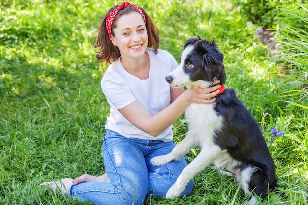 Lächelnde junge attraktive Frau, die mit niedlichem Welpenhunde-Grenzcollie im Sommergarten oder im Stadtparkhintergrund spielt.