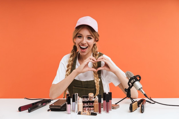Lächelnde junge blonde Bloggerin, die ein Beauty-Video mit Make-up macht, während sie am Tisch sitzt, isoliert auf Rot