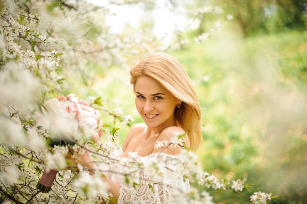 Lächelnde junge blonde Frau im weißen Kleid mit einem Blumenstrauß nahe dem blühenden Kirschbaum