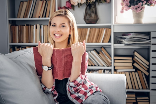 Lächelnde junge blonde Frau in lässiger Kleidung, die ein Buch hält und sich auf einem Trainer in einem Wohnzimmer entspannt.