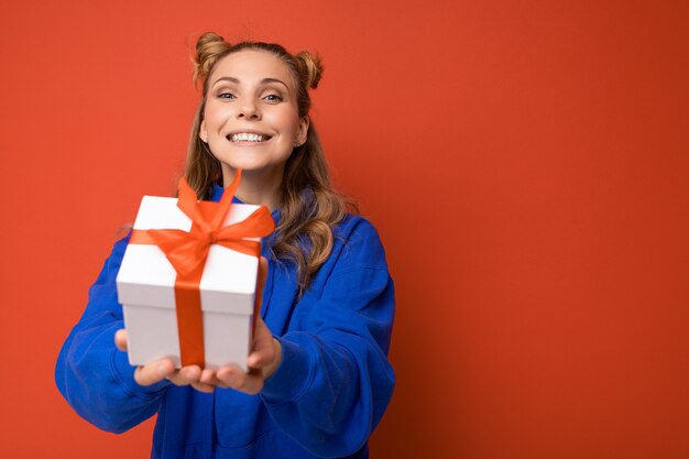 lächelnde junge blonde Frau lokalisiert über bunte Hintergrundwand, die alltägliches trendiges Outfit hält, das Geschenkbox hält und Kamera betrachtet. Speicherplatz kopieren