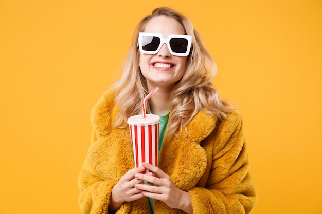 Lächelnde junge blonde Frau Mädchen im gelben Pelzmantel, dunkle Sonnenbrille posiert isoliert auf orangefarbenem Hintergrund im Studio. People-Lifestyle-Konzept. Mock-up-Kopienbereich. Plastikbecher mit Cola oder Soda halten.