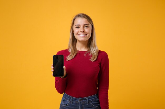 Lächelnde junge blonde Frau Mädchen in Freizeitkleidung posiert isoliert auf gelb-orangeem Hintergrund Studioporträt. People-Lifestyle-Konzept. Mock-up-Kopienbereich. Halten Sie das Mobiltelefon mit einem leeren, leeren Bildschirm.