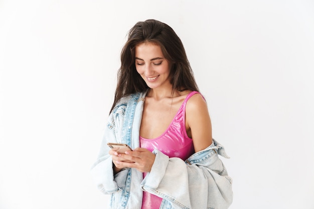 lächelnde junge brünette Frau im Badeanzug stehend isoliert auf weiß, mit Handy