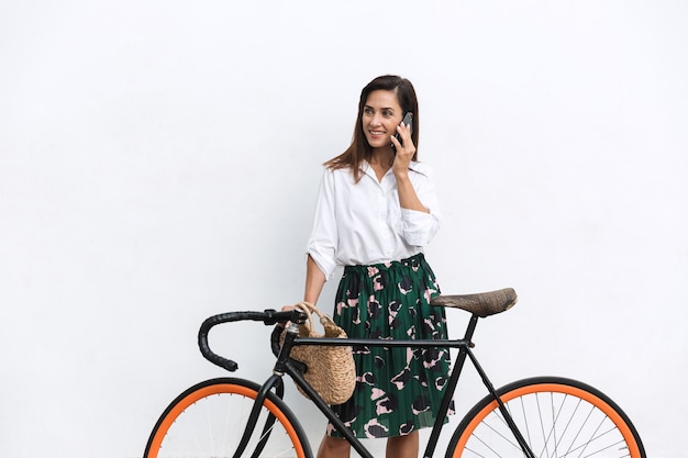 Lächelnde junge brünette Frau in Sommerkleidung, die draußen an der Wand steht, Fahrrad hält und mit dem Handy spricht