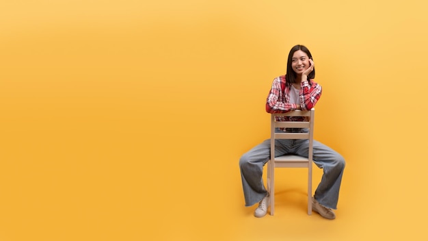 Lächelnde junge chinesische Dame sitzt beiläufig auf einem Stuhl, isoliert auf gelbem Hintergrund, Studio-Panorama