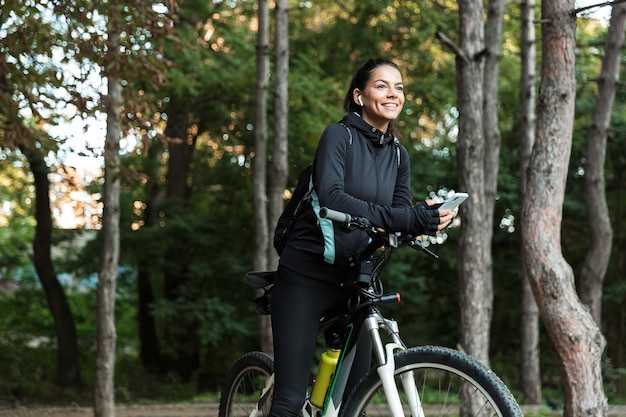 Lächelnde junge Fitnessfrau, die auf einem Fahrrad am Park reitet, Musik mit Kopfhörern hört, Handy hält