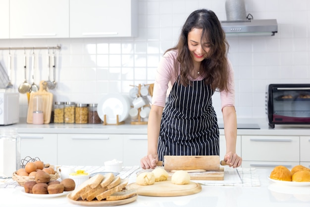 Lächelnde junge Frau arbeitet mit Holzrollenstift, die süßen Kuchen oder Gebäckteig in der Küche herstellt, glückliche tausendjährige Frau, die in der Schürze kocht, die Familienessen oder Dessertbrötchen zubereitet