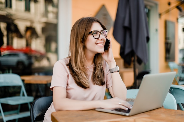 Lächelnde junge Frau, die am Telefon spricht und draußen auf Laptop im Café schreibt