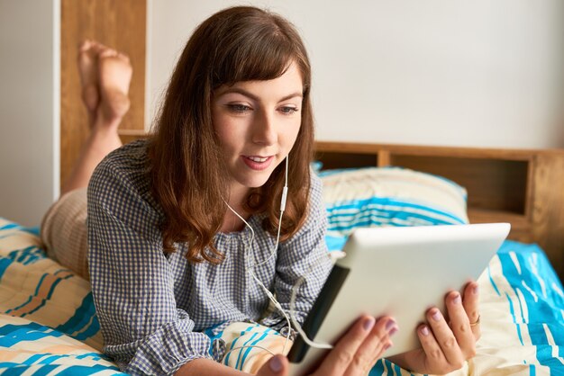 Lächelnde junge Frau, die auf dem Bett liegt und sich eine Show oder Sitcom auf einem digitalen Tablet ansieht?