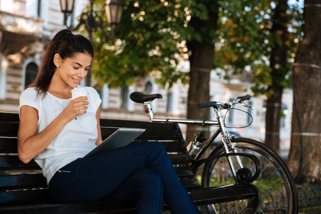 Lächelnde junge Frau, die auf der Bank mit Tasse Kaffee ruht und Tablette im Freien verwendet