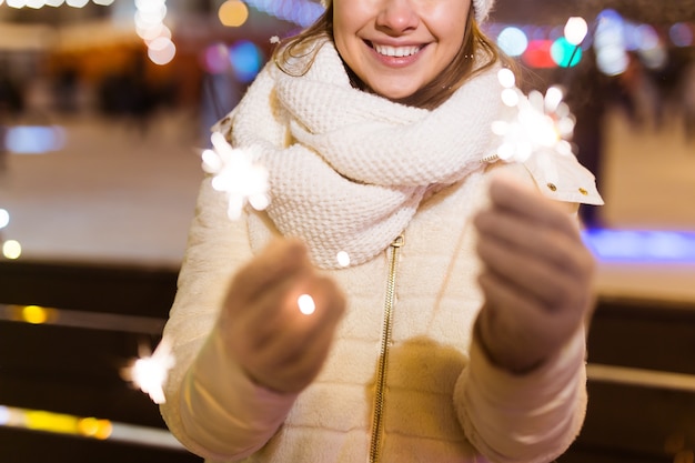 Lächelnde junge Frau, die den Winter trägt, strickte Kleidung, die draußen Wunderkerze über Schneehintergrund hält. Weihnachtsferien.