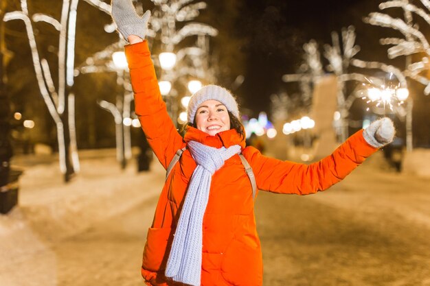 Lächelnde junge frau, die den winter trägt, strickte kleidung, die draußen wunderkerze über schneehintergrund hält. weihnachtsferien.