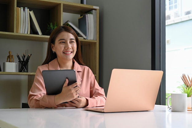 Lächelnde junge Frau, die digitale Tablette hält und am Schreibtisch sitzt.