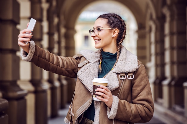 Lächelnde junge Frau, die draußen steht, Einwegbecher mit Kaffee hält und Selfie nimmt.