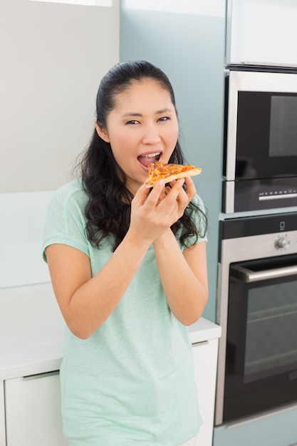 Lächelnde junge Frau, die eine Scheibe der Pizza in der Küche isst