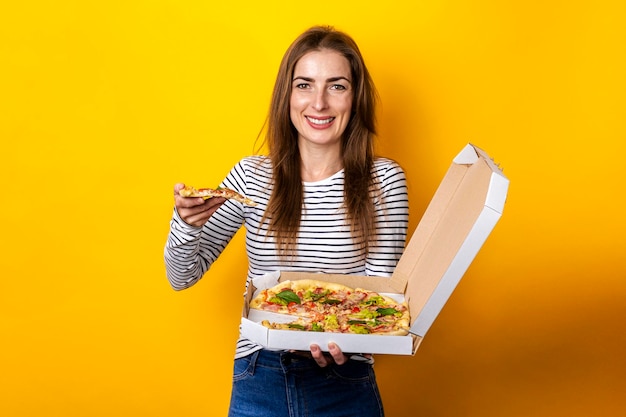 Lächelnde junge Frau, die frische heiße Pizza mit Verpackung auf gelbem Hintergrund isst