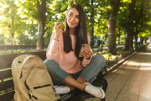 Lächelnde junge Frau, die Handy hält, während sie auf einer Bank im Stadtpark sitzt und Apfel isst