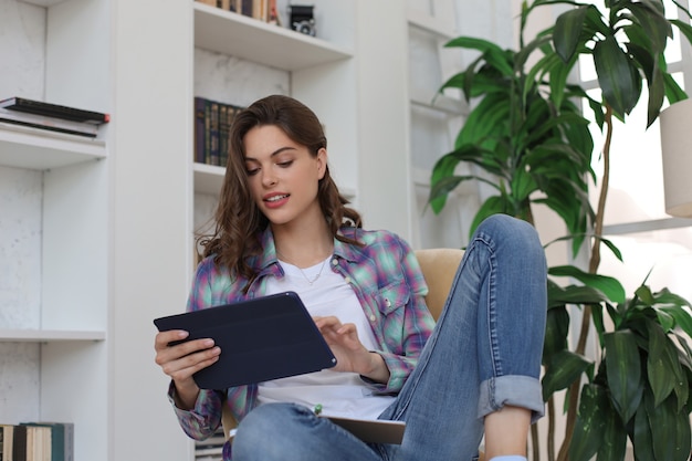 Lächelnde junge Frau, die im Sessel im Wohnzimmer sitzt und mit ihrem Tablet verwendet.