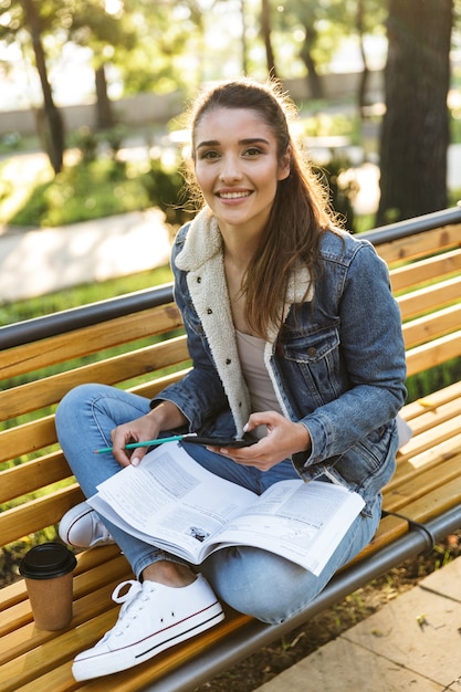 Lächelnde junge Frau, die Jacke trägt, die auf einer Bank im Park sitzt und Zeitschrift liest