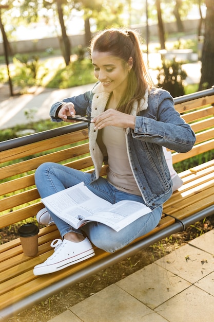 Lächelnde junge Frau, die Jacke trägt, die auf einer Bank im Park sitzt, Zeitschrift liest, ein Foto macht