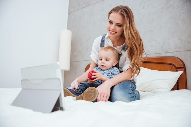 Lächelnde junge Frau, die mit ihrem Sohn auf dem Bett sitzt und Cartoons auf dem Tablet sieht