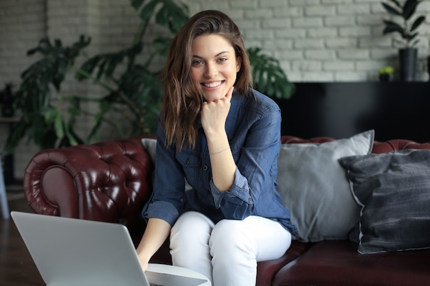 Lächelnde junge Frau, die mit Laptop-Computer auf Sofa sitzt und mit Freunden plaudert.