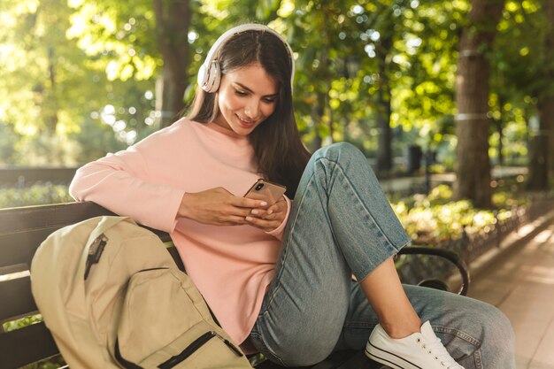 Lächelnde junge Frau, die Musik mit Kopfhörern hört, während sie auf einer Bank im Stadtpark sitzt und Handy hält