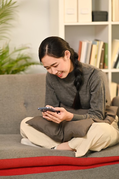 Lächelnde junge Frau, die SMS-Nachrichten schreibt und gerne in sozialen Netzwerken auf dem Smartphone chattet, während sie zu Hause auf der Couch sitzt