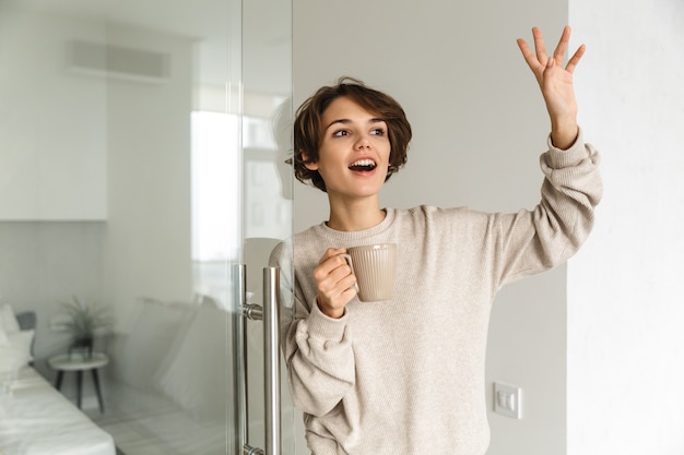 Lächelnde junge Frau, die Tasse hält, während sie am Morgen im Schlafzimmer steht und Hand streckt