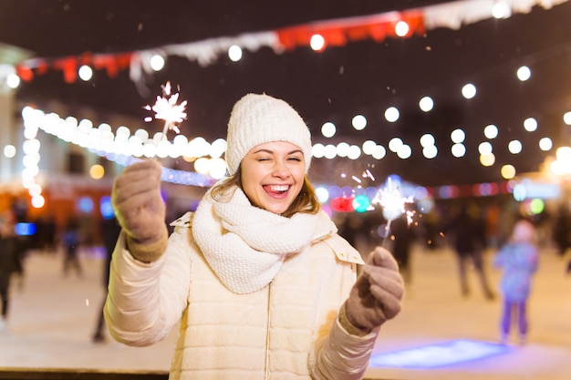 Lächelnde junge Frau, die Winterstrickkleidung trägt, die Wunderkerze im Freien über Schneehintergrund hält