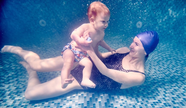 Foto lächelnde junge frau im badeanzug hält ein entzückendes kind in den armen, während sie unter wasser auf dem boden des swimmingpools liegt liebende mutter lehrt ihren sohn, unter wasser zu schwimmen konzept von familie und schwimmen