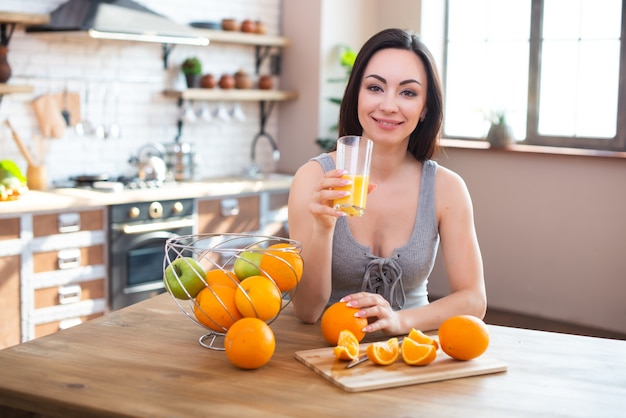 Lächelnde junge Frau im grauen T-Shirt trinkt frischen Orangensaft im Kücheninnenraum