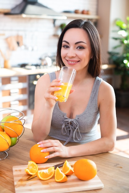 Lächelnde junge Frau im grauen T-Shirt trinkt frischen Orangensaft im Kücheninnenraum