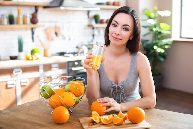 Lächelnde junge Frau im grauen T-Shirt trinkt frischen Orangensaft im Kücheninnenraum