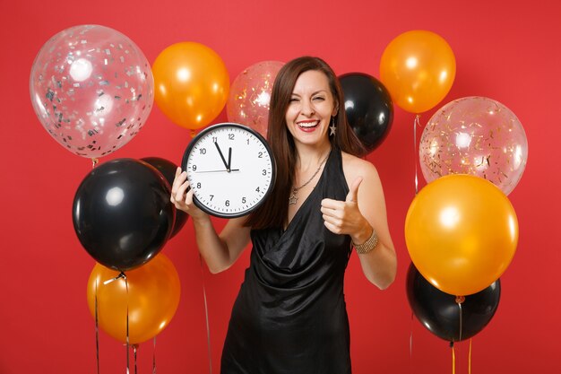 Lächelnde junge Frau im schwarzen Kleid, die das Halten der runden Uhr feiert und Daumen auf hellroten Hintergrundluftballons zeigt. Valentinstag, guten Rutsch ins Neue Jahr, Geburtstagsmodell-Feiertagspartykonzept.