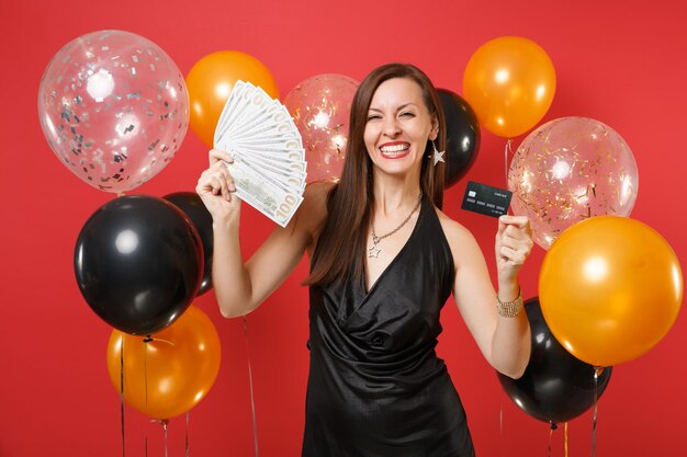 Lächelnde junge Frau im schwarzen Kleid, die Kreditkarte feiert, bündelt viele Dollar Bargeld auf roten Hintergrundluftballons. Valentinstag Happy New Year Geburtstag Mockup Urlaub Party Konzept.