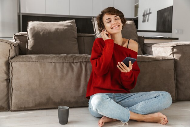 Lächelnde junge Frau in den Kopfhörern, die Pullover tragen, die auf einer Couch zu Hause entspannen